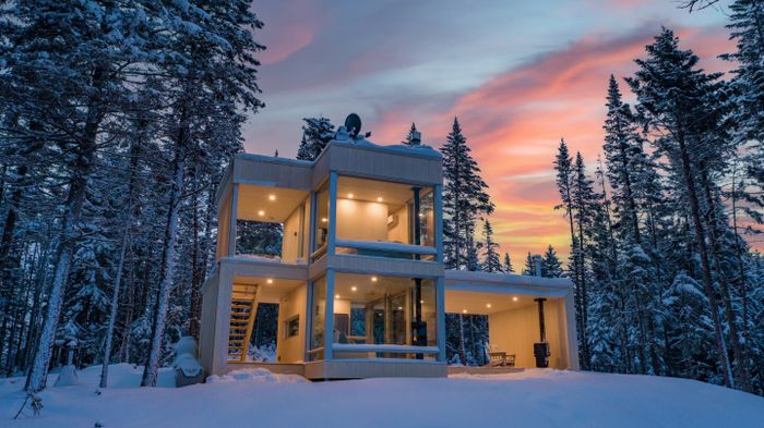 A two-story, minimalist tiny house aglow with lights, surrounded by evergreen trees in a snowy forest under a sky painted with pink and yellow clouds.