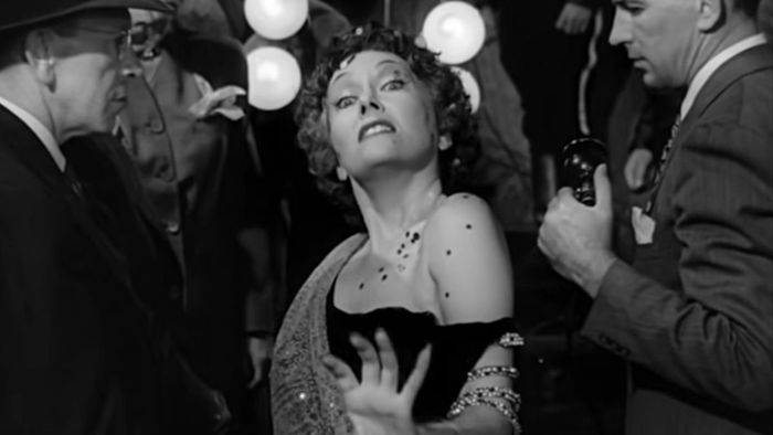 Gloria Swanson, trang điểm và mặc váy, đi trước máy quay của cảnh sát trong Sunset Boulevard