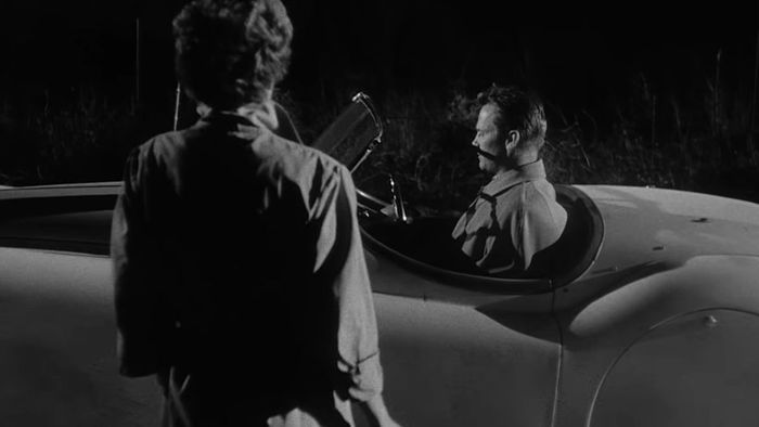 Một phụ nữ tiếp cận một người đàn ông trong một chiếc ô tô convertible trong Kiss Me Deadly