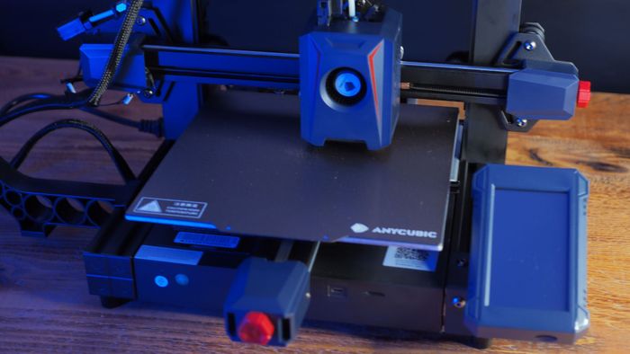 Anycubic Kobra 2 đã được lắp ráp hoàn chỉnh trên một cái bàn gỗ, với phía sau là nền đen