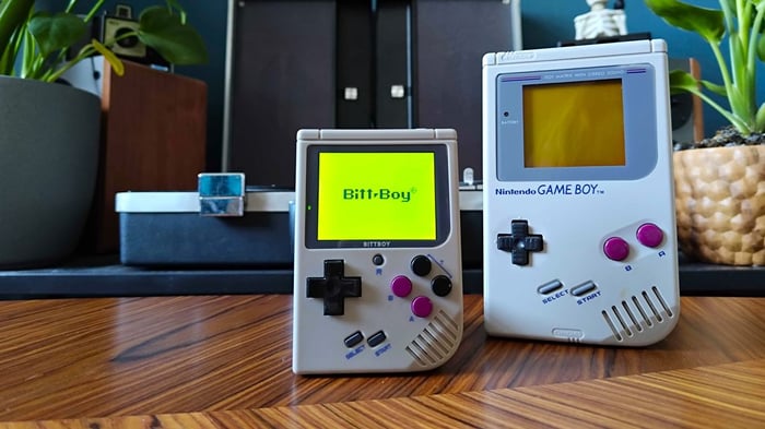 Bittboy cầm tay bên cạnh Game Boy trên bàn màu gỗ