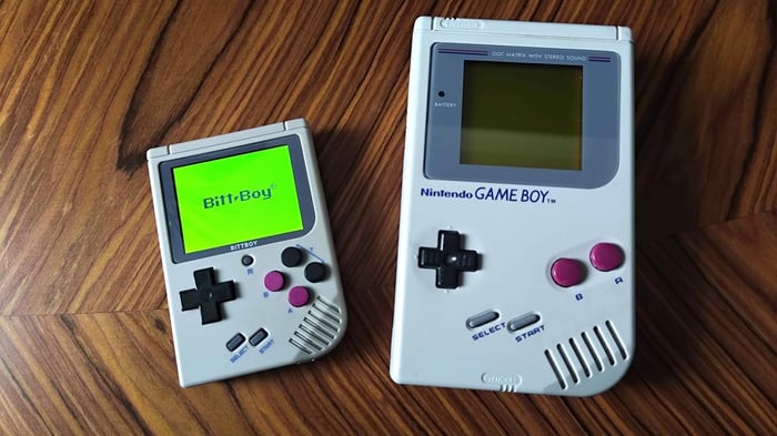 Bittboy bên cạnh Gameboy gốc trên bàn gỗ vân gỗ