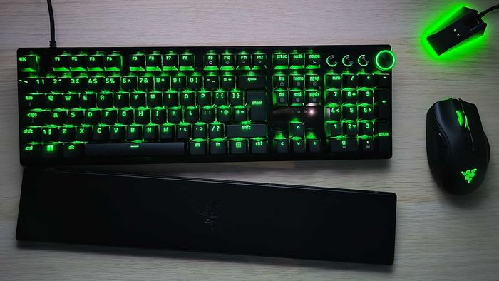 Một chiếc Razer Huntsman V3 Pro nhìn từ trên xuống trên một bề mặt gỗ cùng một chuột và bộ cắm chuột, với bàn đỡ cổ tay hơi cách xa