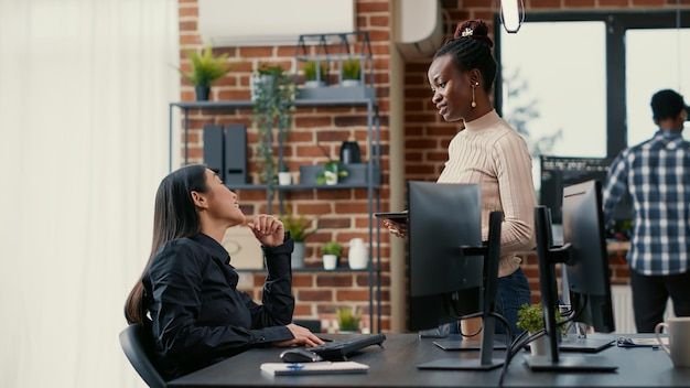 High-Quality Image | Programmer standing and holding digital tablet while conversing with coder seated at desk in front of computer screen displaying running code. Software developer coding user interface for cloud computing online.