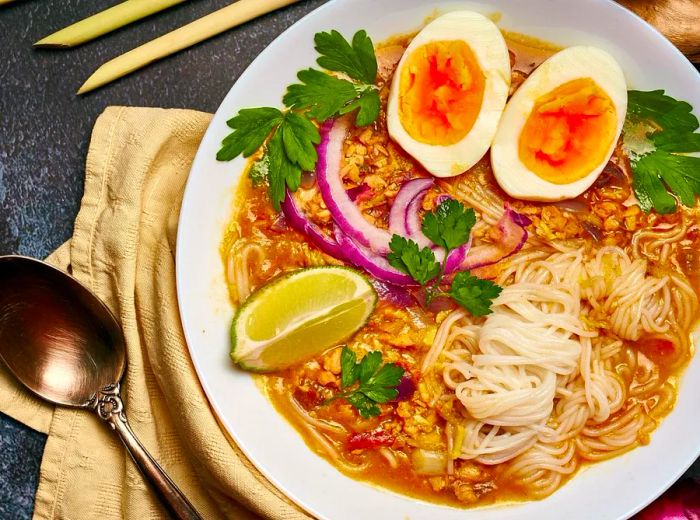 A steaming bowl of noodles topped with a boiled egg, fresh cilantro, sliced onion, and a wedge of lime.