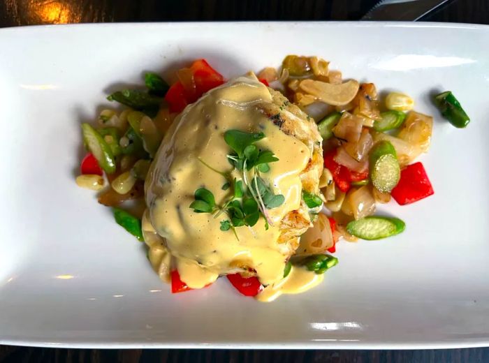 A generous crab cake smothered in savory sauce, resting on a bed of fresh vegetables.