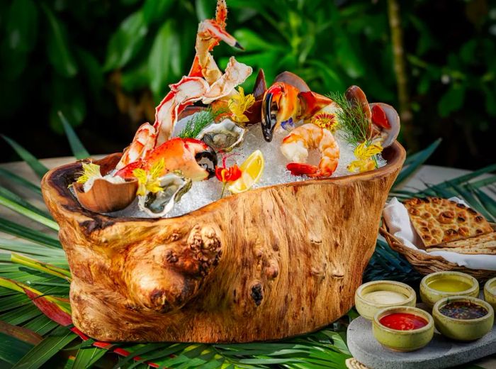 An assortment of raw seafood elegantly displayed on ice within a large tree stump, accompanied by various sauces and condiments on a palm-adorned table.