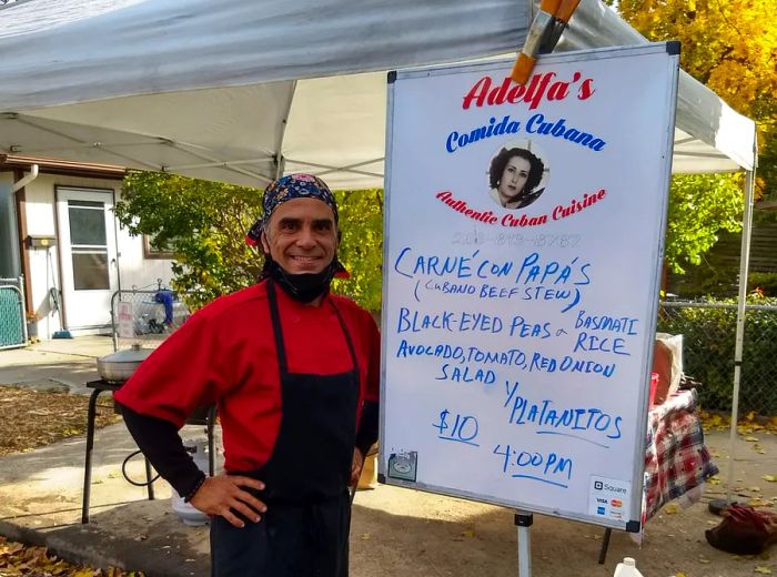 A chef in an apron stands next to a whiteboard adorned with his food stand’s name and a list of available dishes.