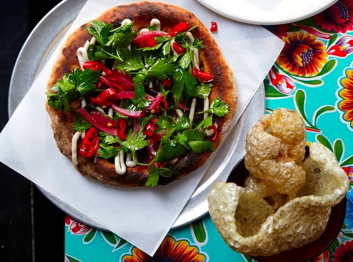 A flatbread loaded with meat and vegetables, complemented by crispy pork crackling, all set on a vibrant tablecloth.