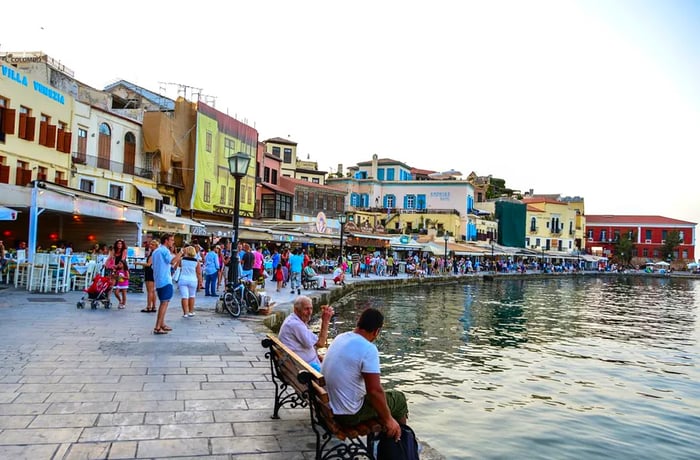 Strollers explore the perimeter of an ancient port, flanked by vibrant restaurants and charming houses.