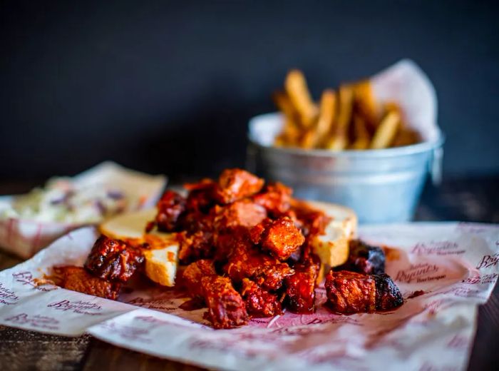 Tender chunks of burnt ends drenched in a deep red barbecue sauce, served atop a slice of soft white bread with a side of crispy fries.