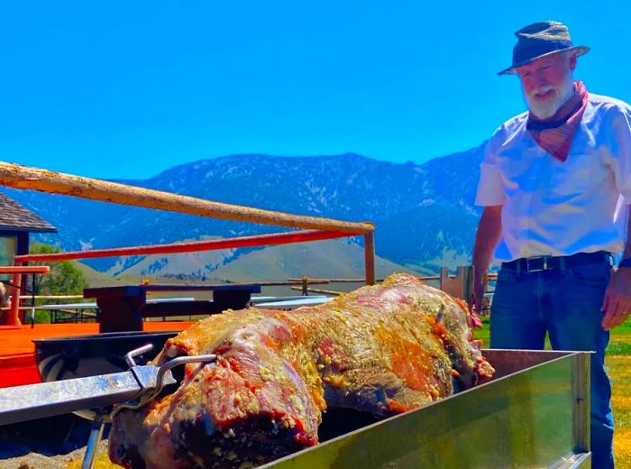 A man oversees a lamb slowly roasting on a spit against a backdrop of a clear blue sky and rugged mountains.