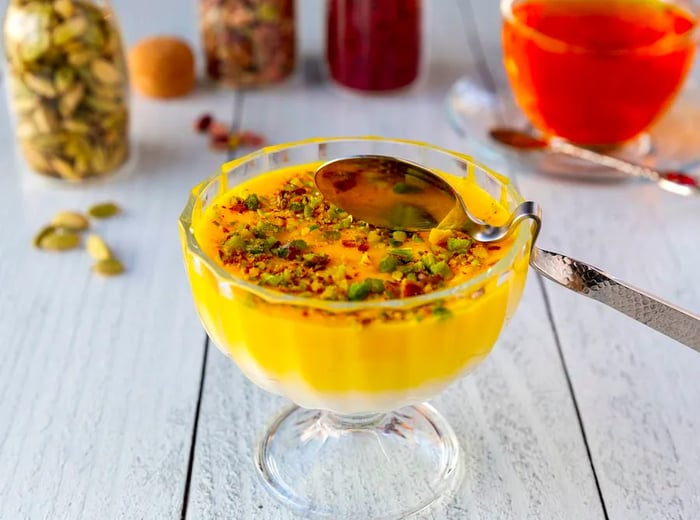 A vibrant yellow pudding adorned with chopped pistachios, served with a uniquely curved spoon designed to rest over the rim of the dish.