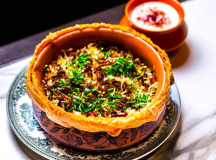 A vibrant pot of biryani with a doughy crust lifted to reveal the colorful layers inside.