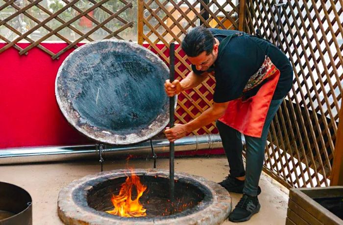 A chef skillfully stirs a fiery pit embedded in the ground using a long pole.