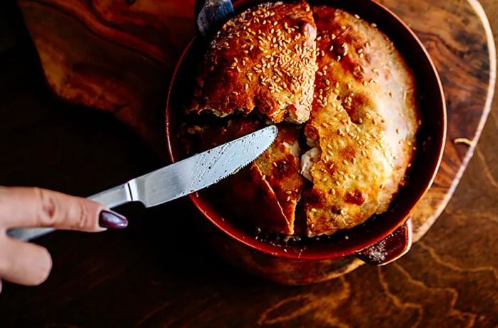 A hand holding a knife cuts into a meat pie, which is served in a skillet.