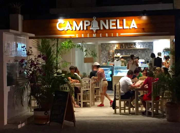 In the evening, patrons enjoy their time at outdoor tables illuminated by the glowing Campanella Cremerie sign. The gelateria’s interior is visible through large windows, showcasing the bustling ice cream counter and friendly staff serving up delightful treats.