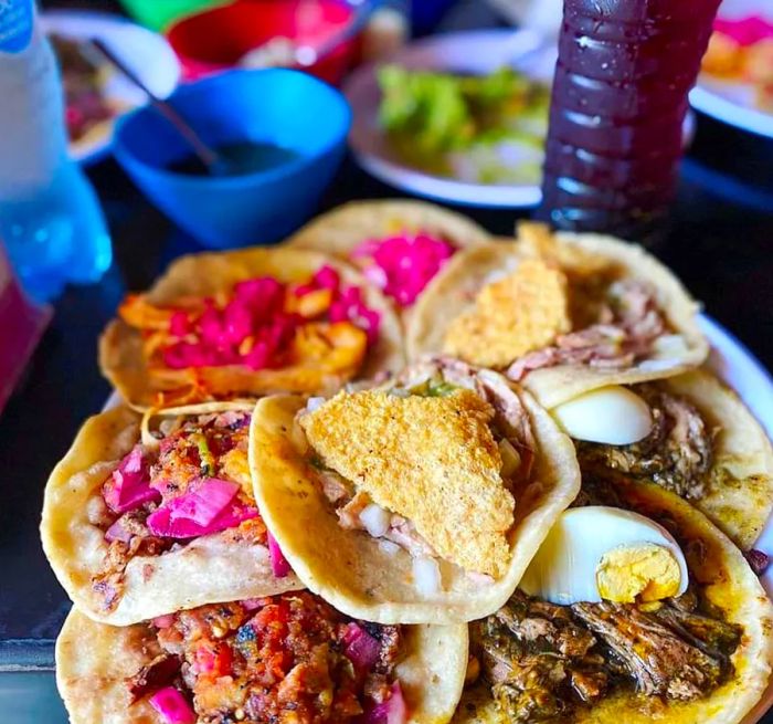 A selection of tacos presented on a tray, featuring various ingredients like chicharron, boiled egg, meat, and pickled vegetables.