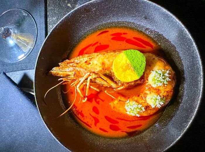 A whole prawn swimming in a red broth, garnished with dollops of emulsion and a lemon wedge sprinkled with green powder.