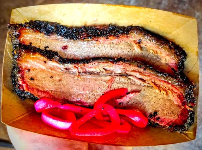 Two slices of brisket served in a cardboard container, garnished with vibrant pink pickled onions.