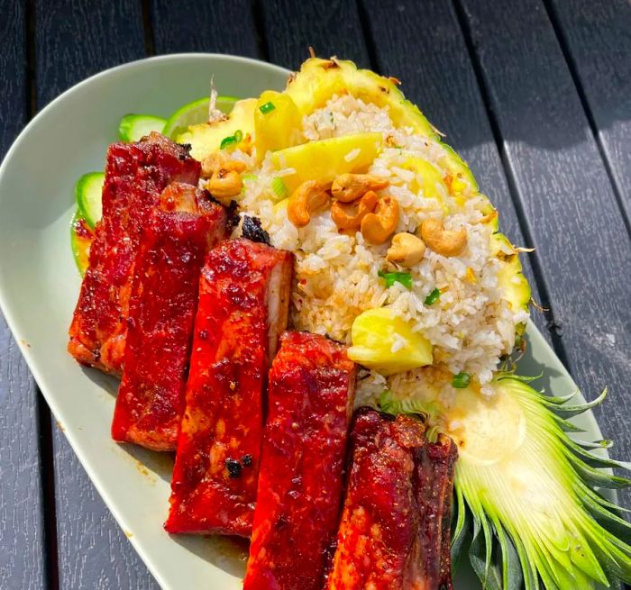 A serving of saucy pork ribs resting against a pineapple half filled with fried rice.