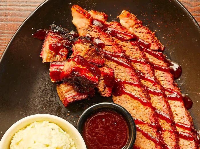 A generous serving of burnt ends, brisket slices bathed in sauce, accompanied by ramekins of savory sides on a plate.