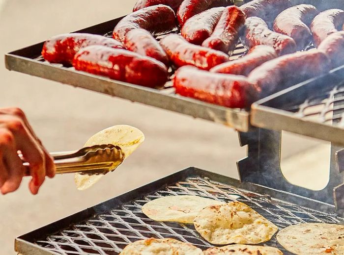 A chef maneuvers a tortilla with tongs on one rack of a grill, while sausages sizzle on another.