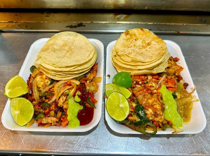 Two trays of tacos, each with stacked tortillas in the back and a variety of ingredients arranged in the front, set on a metal counter.