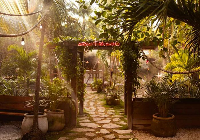 A vibrant entrance to an al fresco dining area, surrounded by dense greenery with sunlight filtering through the leaves, and a glowing neon sign reading Gitano.