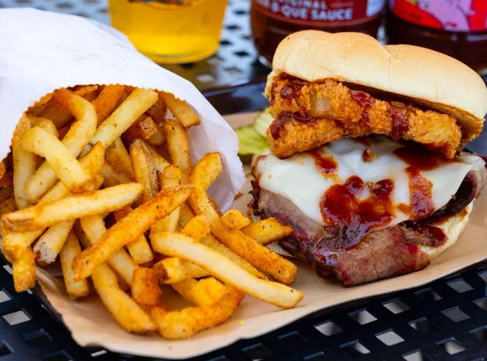 A savory sandwich featuring brisket smothered in melted cheese and topped with crispy onion rings, resting on a napkin beside a generous serving of golden French fries.