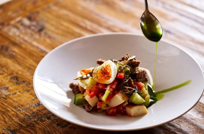 A spoon gently drizzles oil over a salad composed of chopped vegetables and boiled egg.