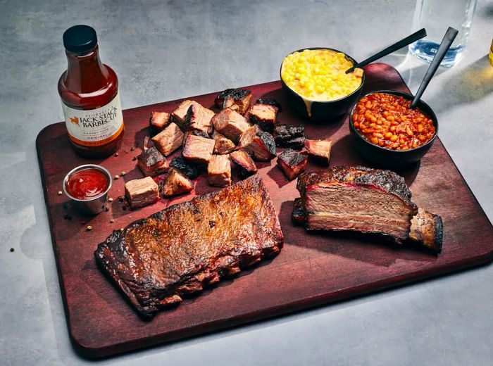 A wooden platter piled high with succulent ribs, juicy burnt ends, creamy mac and cheese, and hearty baked beans, accompanied by a bottle of tangy barbecue sauce.