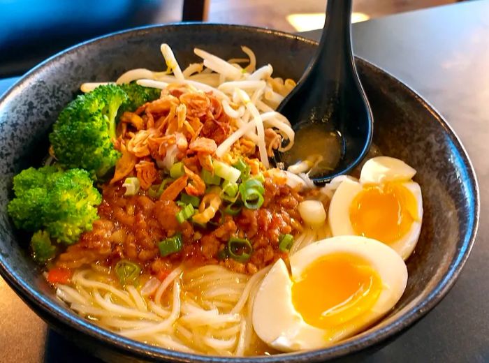 A bowl of Shan noodles garnished with a boiled egg, broccoli, bean sprouts, and tender meat.