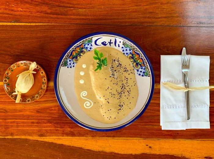 A top-down view of a fish fillet on a branded plate, covered in a rich white mole sauce, garnished with a sprig of parsley, dark chocolate drizzle, and sesame seeds, placed on a wooden table beside serveware and a small package wrapped in corn husk.