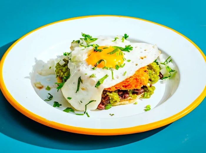 A breakfast taco featuring beans, potatoes, and a fried egg, presented on a white plate with a yellow rim against a blue background.