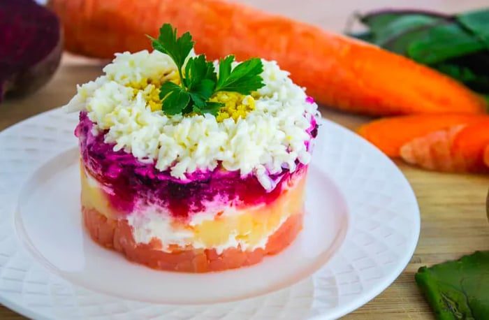 A plate featuring layers of salmon, potatoes, carrots, and beets molded into a cylinder, alongside raw whole carrots on a wooden countertop.