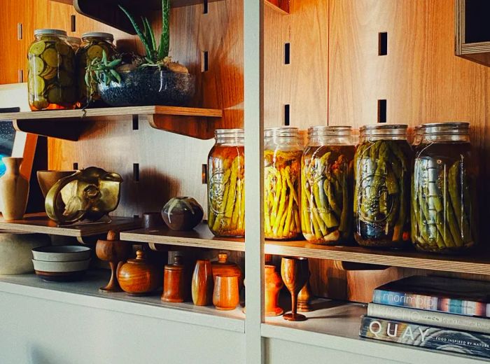 Shelves adorned with jars of vibrant vegetable pickles, accompanied by wooden serveware, artwork, and cookbooks.