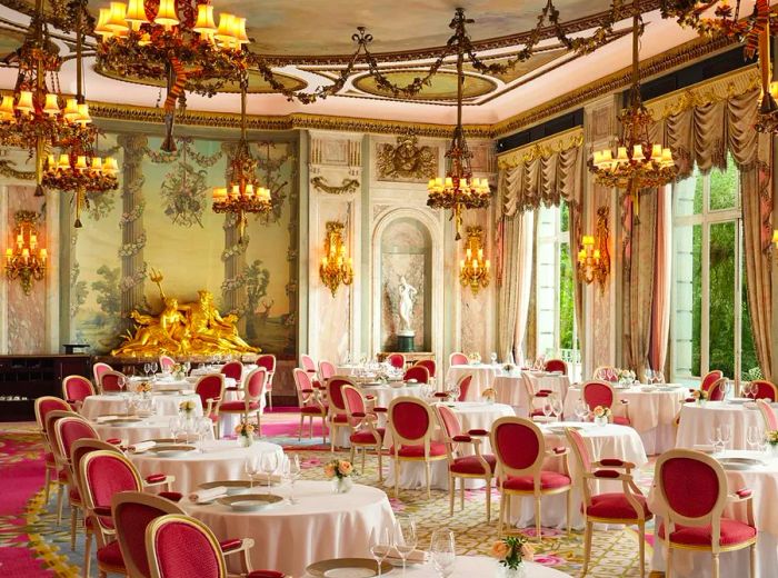 An extravagant dining room adorned with chandeliers, gilded details, expansive windows, and tables draped in crisp white tablecloths.