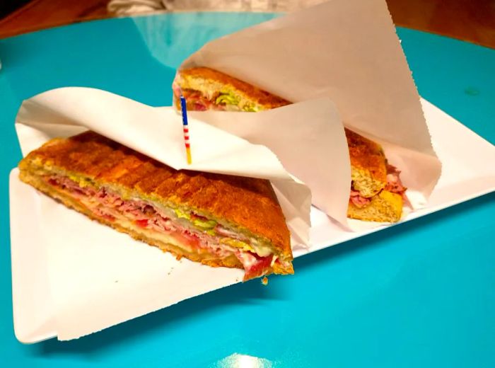 A panini, adorned with an American flag toothpick, cut into halves and wrapped in butcher paper, rests on a vibrant blue table.