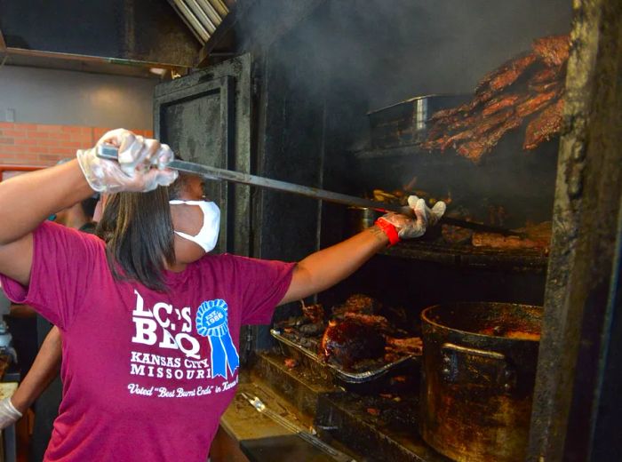 A staff member in a branded t-shirt skillfully adjusts meats in a massive smoker using a long-handled tool.