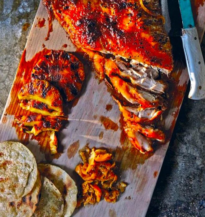 A large slab of roast pork belly, served with slices, alongside roasted pineapple and tortillas.