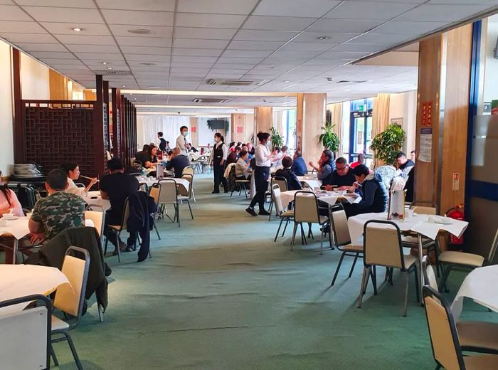 A spacious dining area featuring a green carpet, large round tables, and diners enjoying their meals while waitstaff move around taking orders.