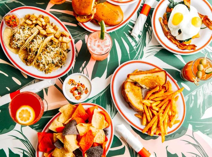 Various brunch dishes including sandwiches with fries, tacos, eggs, and tortilla chips served on a tropical-patterned table.