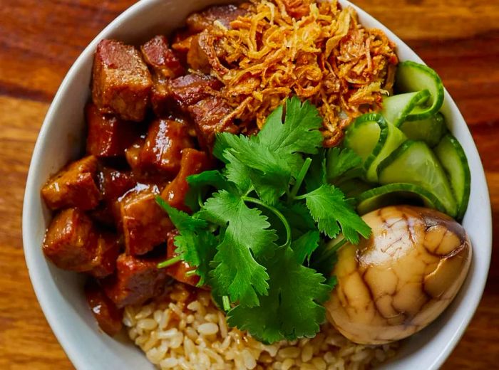 A bowl of lu rou fan featuring tender chopped pork belly, tangy pickled cucumber, crispy fried shallots, fresh cilantro, a tea egg, and a bed of rice.