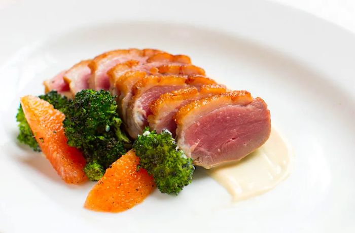 A plate of rare-sliced duck breast served with broccoli and orange wedges.