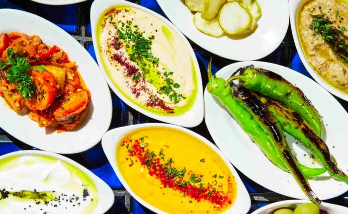 An overhead view of a table loaded with vibrant dishes, including grilled peppers, various dips, and pickles.