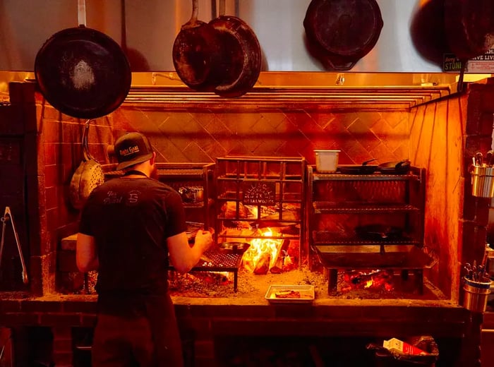 A chef stands before a large heart-shaped setup, surrounded by metal racks and a roaring fire in the center.