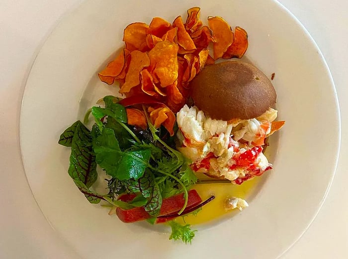 A delectable roll filled with chopped lobster and crab, served on a plate alongside sweet potato chips and greens, all set on a white tabletop.