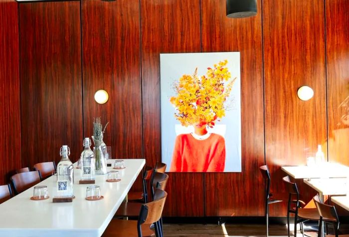 A wood-clad dining area featuring long white tables with water pitchers, adorned with a striking piece of art depicting a person with a head full of blooming flowers.