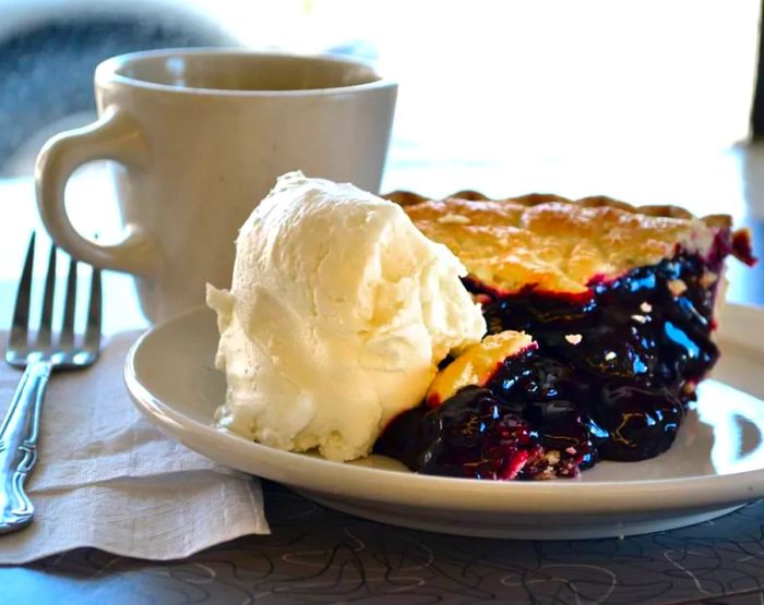 A slice of blueberry or blackberry pie topped with a generous scoop of ice cream, set on a sunny countertop next to a coffee mug and place setting.